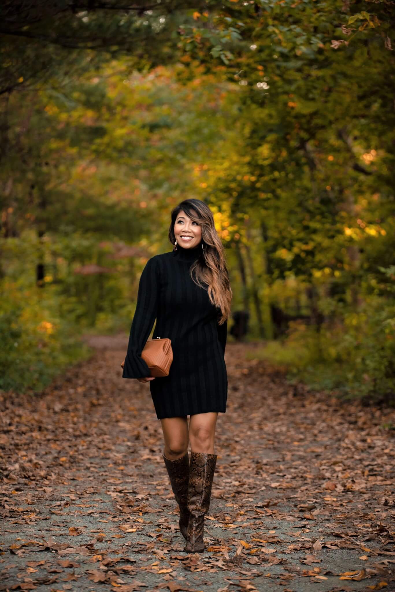 lady with long dark hair in black sweater dress with snakeskin boots and brown clutch walking through the forest during fall