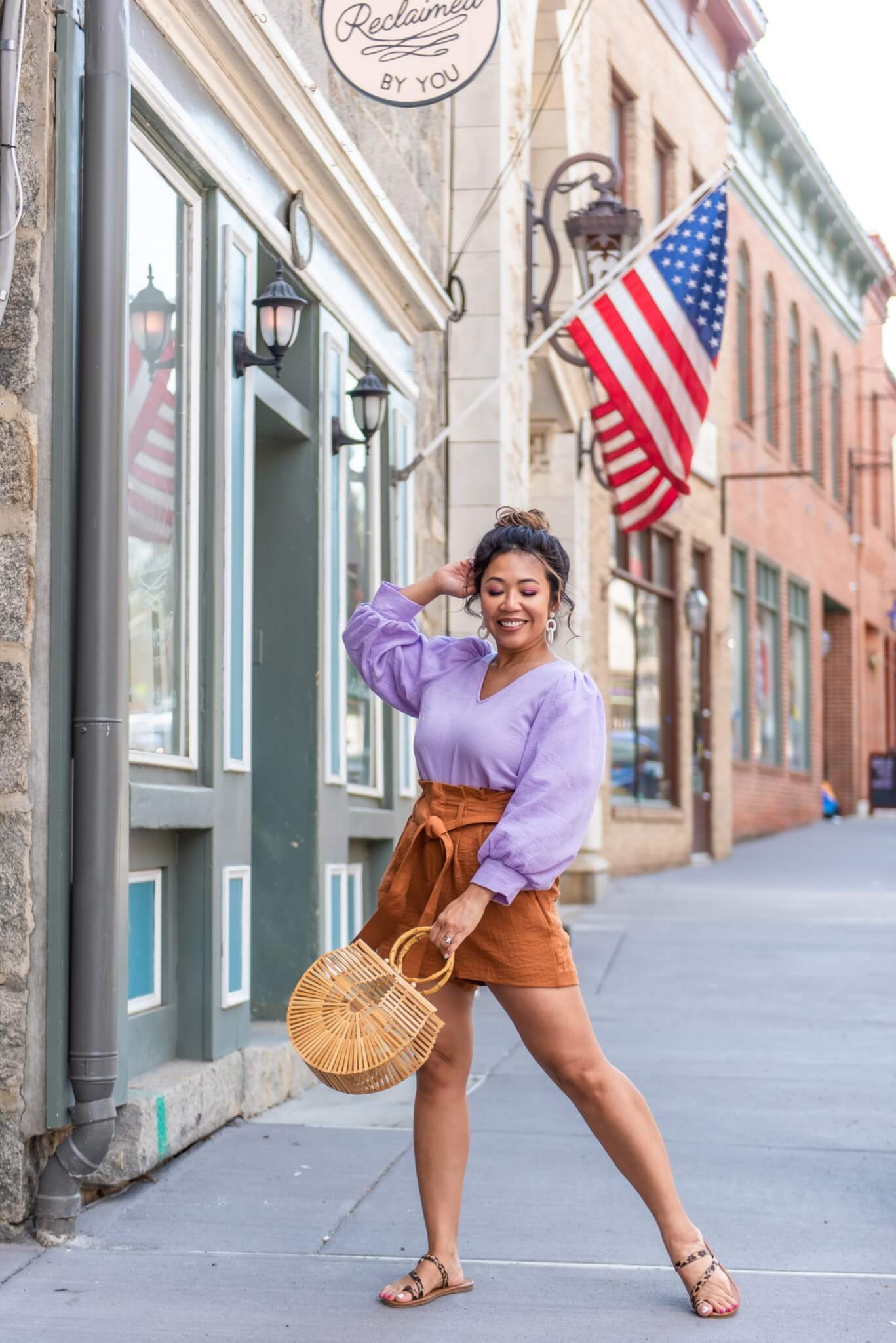 lady posing with one hand up and the other holding a bamboo clutch bag. She's standing in front of the American flag, which is above her head.