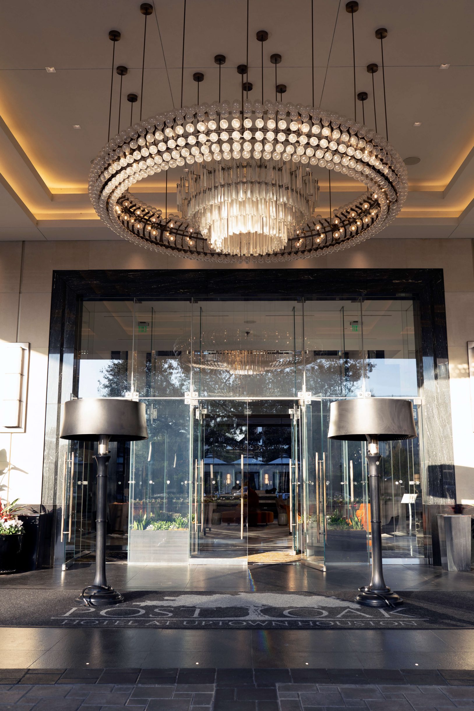 entrance of the post oak hotel at uptown in houston, texas. It has an enourmous circular chandelier, with a rug that has post oak hotel written on it with their emblem (an oak tree). there are two large lampshade lighting flanked to the side.