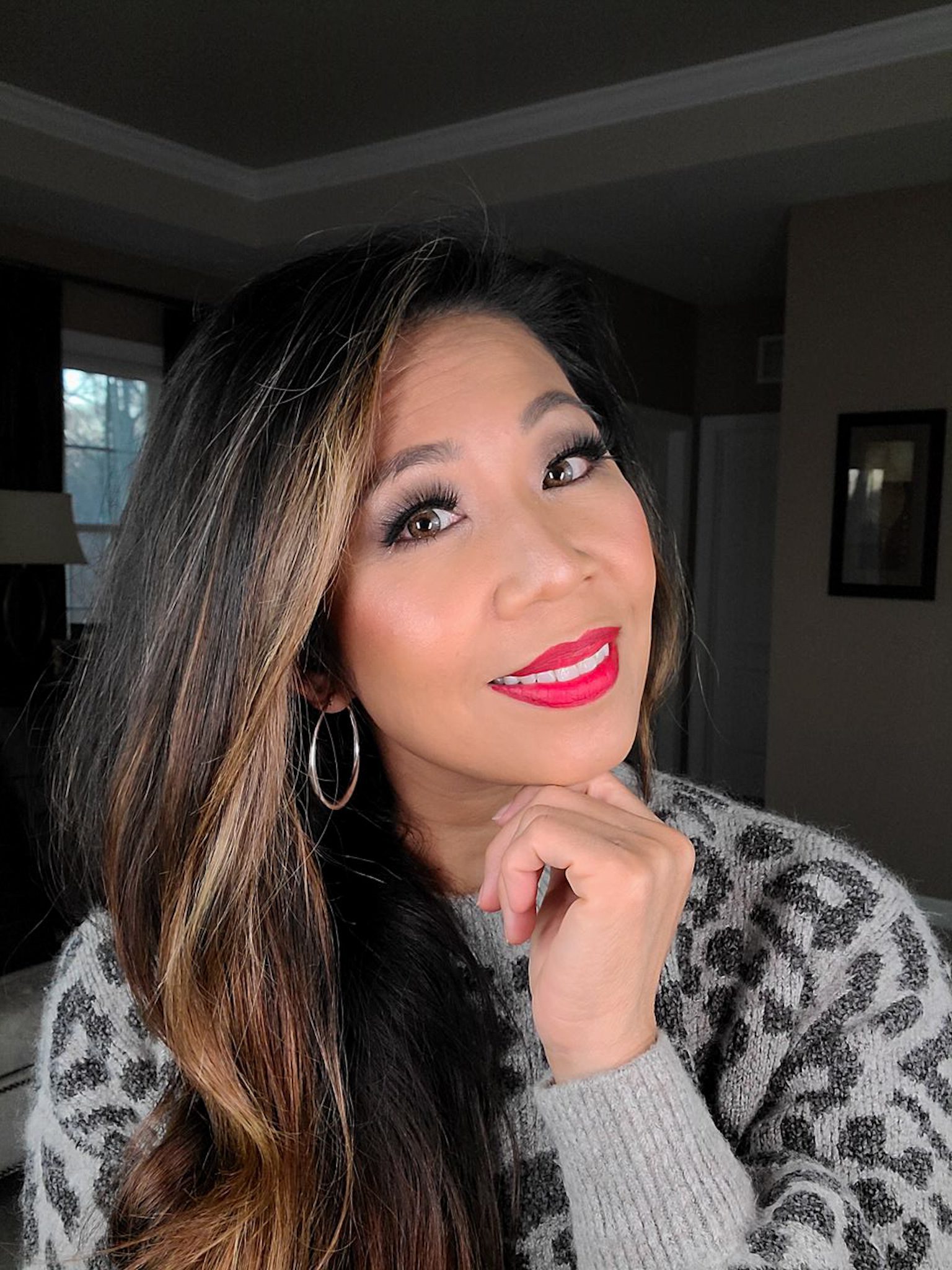 woman smiling with her head slightly tilted to one side and her hand on her chin. she's wearing a gray sweater, smoky gray eyeshadow, and red lipstick.