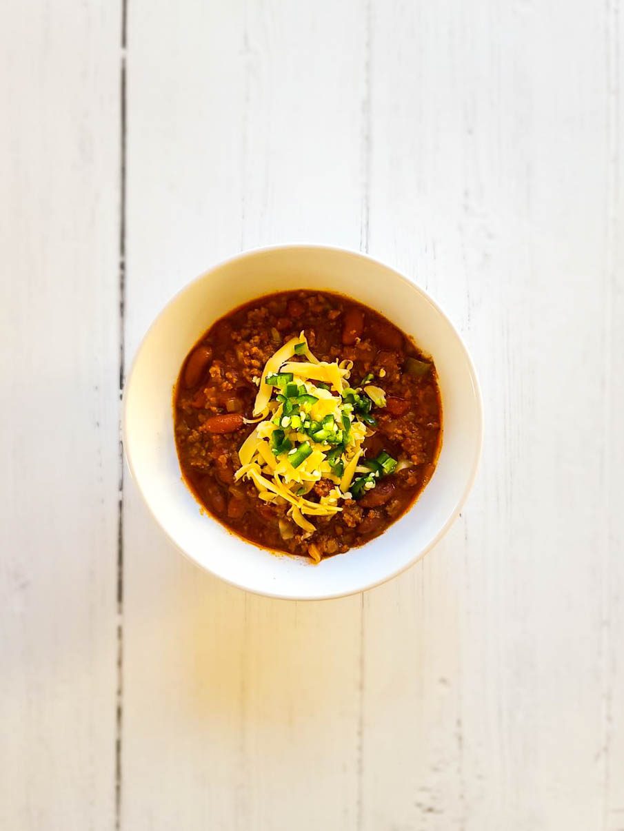 spicy beef chili with beans in a white bowl topped with shredded cheddar cheese and diced alapeno peppers