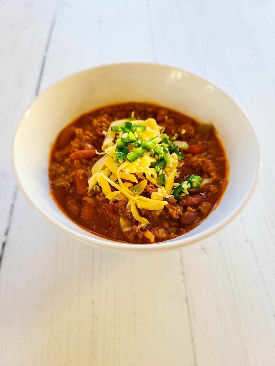 spicy beef chili with beans in a white bowl topped with shredded cheddar cheese and diced alapeno peppers