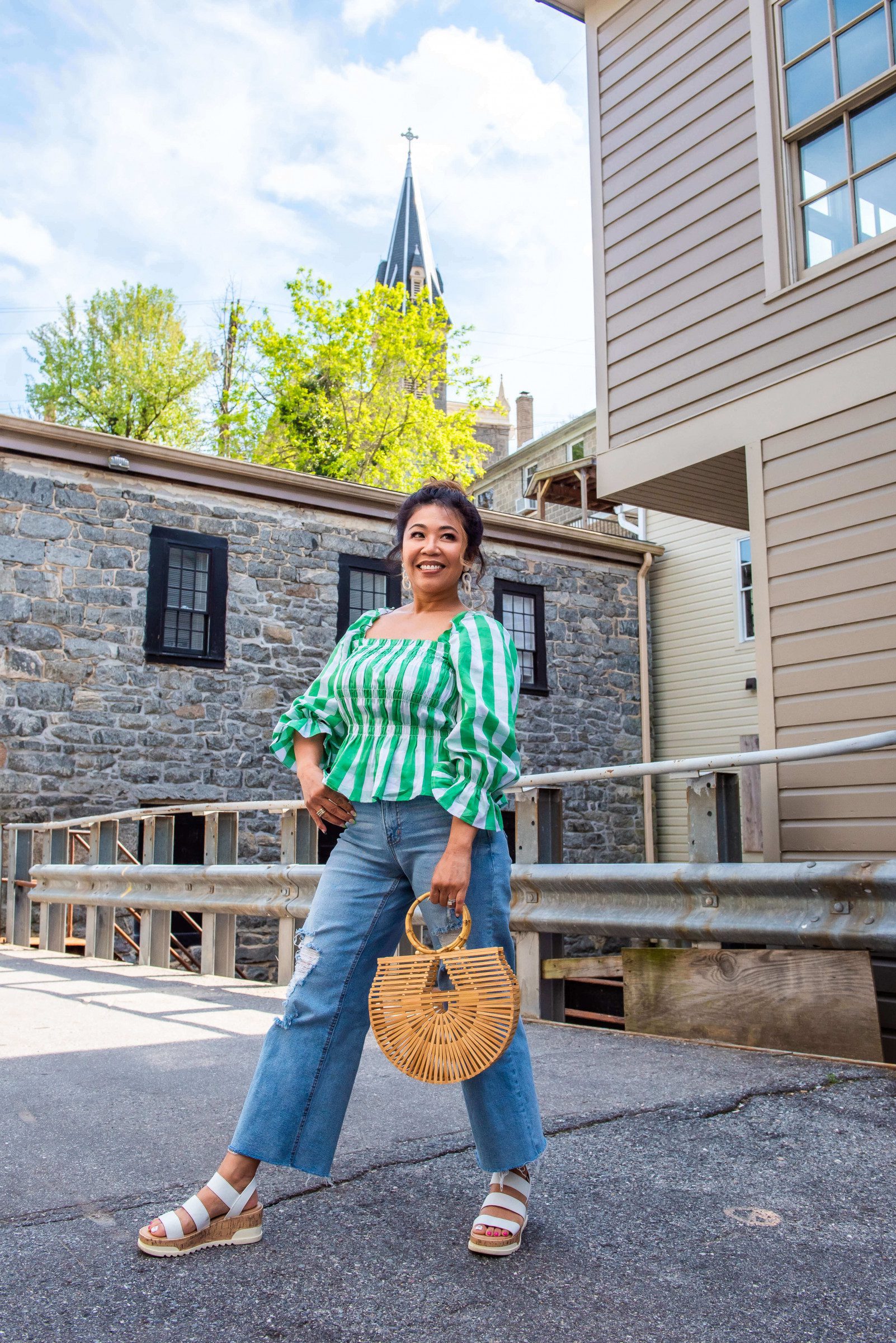 lady wearing a green and white gingham long-sleeve top with deconstructed denim jeans with wicker bag and white sandals.